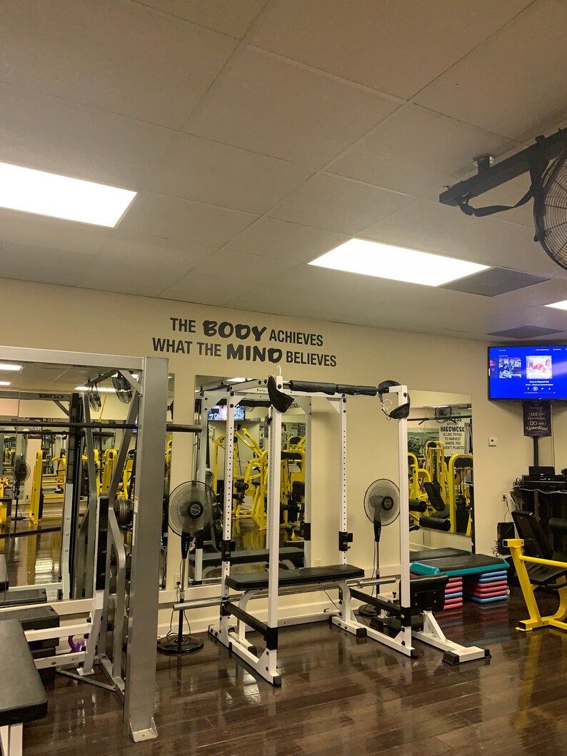 May include: A gym with a white and black weight rack, a mirror, and a motivational quote on the wall that reads "The body achieves what the mind believes."