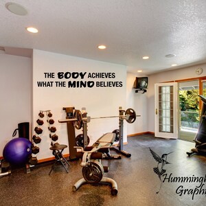 May include: Black and white motivational quote on a white wall in a home gym: "The body achieves what the mind believes". The gym has a weight bench, dumbbells, a treadmill, and a yoga ball.