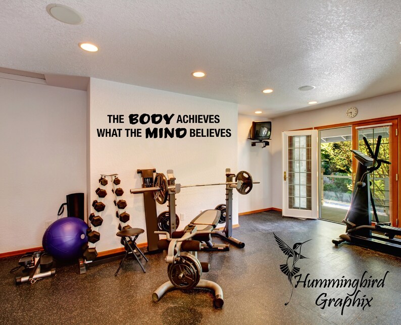 May include: Black and white motivational quote on a white wall in a home gym: "The body achieves what the mind believes". The gym has a weight bench, dumbbells, a treadmill, and a yoga ball.
