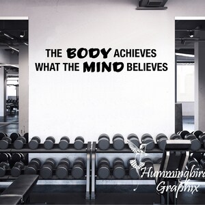 May include: Black and white motivational quote on a white wall in a gym setting. The quote reads "The body achieves what the mind believes".