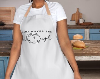 May include: White apron with the text "MAMA MAKES THE DOUGH" in black script. The apron has a tan neck strap and is worn over a white t-shirt. The image is set in a kitchen with wooden countertops and blue cabinets.