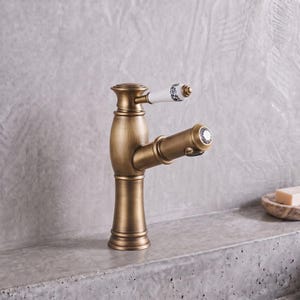 May include: A brass-toned bathroom faucet with a cylindrical base and a curved spout. The handle is white with black floral patterns and a metallic tip. The spout features a circular crystal-like accent at the end, providing a distinct look.