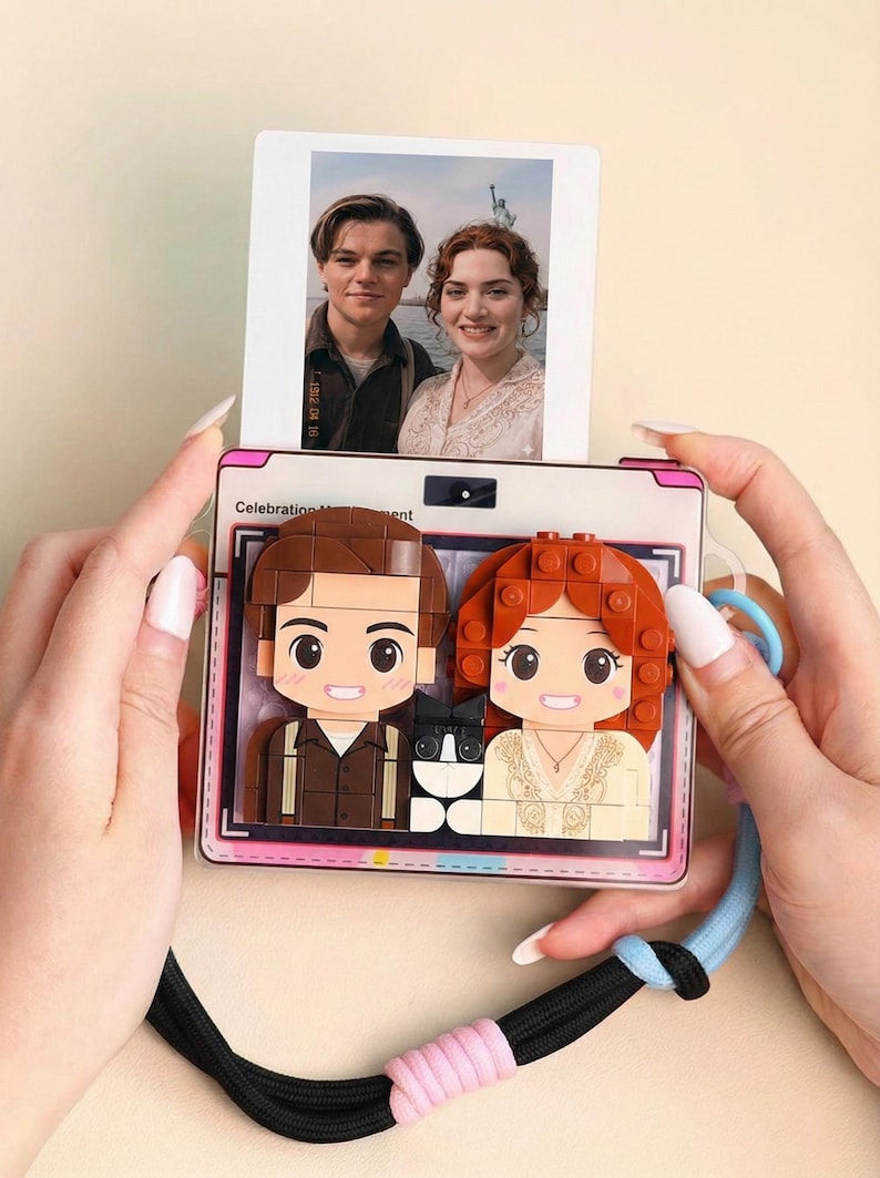May include: A novelty camera-shaped phone case featuring a photo of a couple and a Lego-style design of the same couple with a cat. The case has a pink border and a black and blue strap.