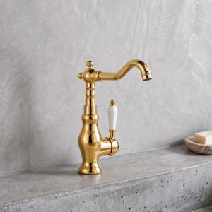 May include: A polished gold-tone metal faucet featuring a curved spout and a single side-mounted handle with a smooth white ceramic accent. The fixture has a classic, ornate base design with tiered detailing and a reflective metallic finish.