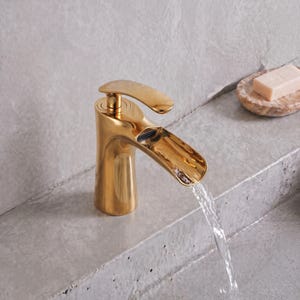 Könnte beinhalten: Eine polierte goldfarbene Badezimmerarmatur mit breitem, flachem Auslauf. Wasser fließt stetig aus dem offenen Auslauf in ein Becken. Ein kleines, rechteckiges Stück Seife liegt auf einer strukturierten, runden Steinschale und spiegelt den Glanz.