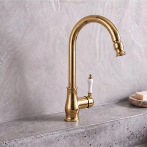 May include: A polished gold kitchen faucet with a high-arc spout and a single side-mounted handle featuring a white ceramic accent. The faucet has a smooth, reflective metallic finish and a curved design that extends upward and then downward.
