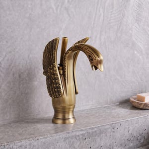 May include: A brass-toned metal tap shaped like a swan with wings extended upwards. The tap features a curved neck, a detailed head, and textured wing feathers. It has a cylindrical base and a vertical handle lever positioned behind the swan neck.
