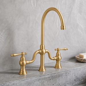 May include: A bridge-style kitchen tap with a brushed gold finish, featuring a high-arc swivel spout and two lever-style handles. The tap has a central pillar design with curved side pipes connecting to the handles on each side.