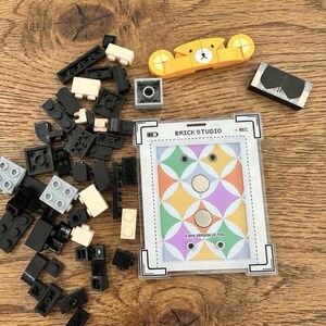 May include: A collection of colorful building blocks and a small yellow bear-shaped toy are scattered on a wooden surface. A card with the text "BRICK STUDIO" and "A MINI VERSION OF YOU" is also present, along with other small toy pieces.