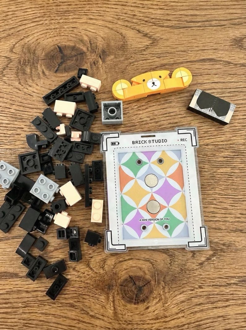 May include: A collection of colorful building blocks and a small yellow bear-shaped toy are scattered on a wooden surface. A card with the text "BRICK STUDIO" and "A MINI VERSION OF YOU" is also present, along with other small toy pieces.