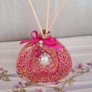 Peut inclure: Un diffuseur rose et doré avec des bâtonnets en bois. La bouteille présente un motif de filigrane doré et est ornée d'un ruban rose, d'une perle et d'un bijou. Le diffuseur est conçu pour contenir des huiles essentielles.