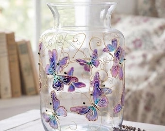 Puede incluir: Un florero de vidrio transparente con mariposas pintadas a mano en tonos morados y azules, decorado con delicadas líneas doradas en espiral. El recipiente cilíndrico tiene un borde ligeramente ensanchado y una superficie lisa y diáfana.