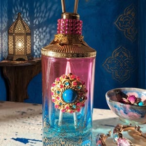 Peut inclure: Un flacon diffuseur orné et coloré, avec un dessus doré et rose, un ornement central en forme de bijou et des bâtonnets en bois. La bouteille passe du bleu au rose, avec des détails en filigrane doré. Un bol décoratif et un bracelet sont en arrière-plan.