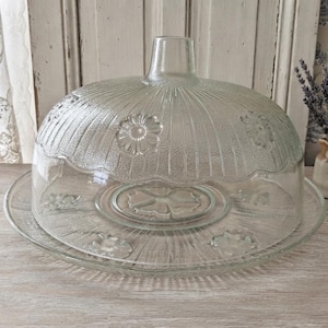 Peut inclure: Un dôme à gâteau en verre transparent avec son plateau assorti, orné d'un motif floral en relief et de stries verticales. Le dôme est surmonté d'un bouton central et le plateau présente un large bord plat. L'ensemble est en verre clair.