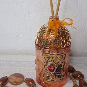 May include: A decorative diffuser bottle with a honeycomb-patterned top and a key charm. The bottle is embellished with jewels and the words "ENJOY LIFE". A necklace with large, oval-shaped beads is in the foreground.