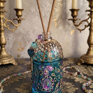 Pode incluir: Um frasco difusor de vidro azul-petróleo com uma tampa decorativa dourada em favo de mel e enfeites de joias ornamentados. Um pequeno pingente de chave e palhetas de madeira são inseridos na garrafa. A garrafa é cercada por castiçais dourados e um colar de contas.