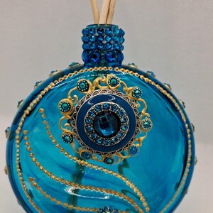 Puede incluir: Un frasco difusor de vidrio azul turquesa con detalles decorativos dorados y azules. La botella presenta una parte superior enjoyada y un medallón central con una gran gema azul. Se insertan cañas de madera en la botella.