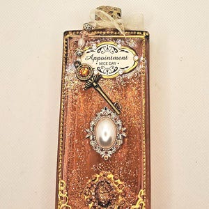 May include: A rectangular, decorative bottle with a bronze-colored glitter interior. Embellishments include a key, a pearl-centered brooch, and a label that reads "Appointment NICE DAY." The bottle is trimmed with gold accents and a ribbon.