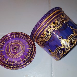 Peut inclure: Un pot en verre violet avec un couvercle, tous deux décorés de peinture dorée et de strass violets. Le pot a un motif de mandala.