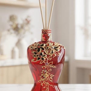 Peut inclure: Un flacon diffuseur à roseaux en verre rouge en forme de torse humain, orné de filigranes floraux dorés, de petits cristaux rouges et d'une gemme rouge en forme de cœur au col. Trois bâtonnets en bois clair sortent de l'ouverture supérieure.
