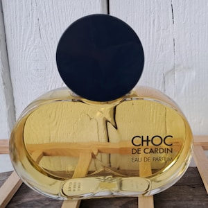 May include: A clear glass bottle of Choc de Cardin Eau de Parfum. The bottle has a round, gold-coloured body and a black, round cap.