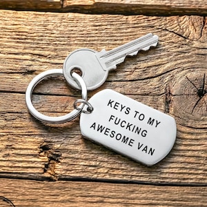 Pode incluir: Uma chave prateada e um porta-chaves com uma etiqueta retangular que diz "KEYS TO MY FUCKING AWESOME VAN" em texto preto. Os itens estão em uma superfície de madeira.