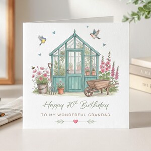 Könnte beinhalten: Quadratische Geburtstagskarte mit einer Aquarellillustration eines türkisgrünen Gewächshauses, Blumen und einer Schubkarre. Der Text lautet: „Alles Gute zum 70. Geburtstag an meinen wunderbaren Großvater“.