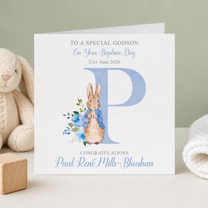 May include: A baptism card with the text "TO A SPECIAL GODSON On Your Baptism Day 21st June 2026". The card features a watercolor illustration of a rabbit wearing a blue jacket, a large blue letter "P", and the name "Paul René Mills-Bhushan".
