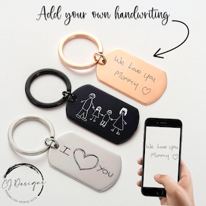 Könnte beinhalten: Personalisierte Schlüsselanhänger in Schwarz, Silber und Roségold mit handgeschriebenen Nachrichten und Familienzeichnungen. Die Schlüsselanhänger haben einen Metallring. Das Bild zeigt auch ein Telefon mit dem Text "We Love you Mommy".