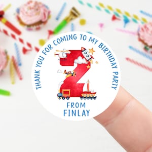 May include: A round white sticker featuring a red number 2 with transport illustrations. The text on the sticker says "Thank you for coming to my birthday party" and "From Finlay". Party theme.