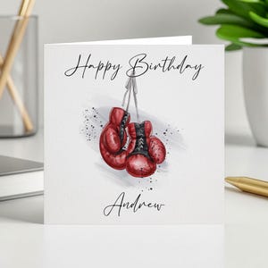 Op de afbeelding: Een wenskaart met de tekst "Happy Birthday" en "Andrew" in sierlijke letters. De kaart toont een aquarel illustratie van rode bokshandschoenen die aan een touwtje hangen, met zwarte spetters.