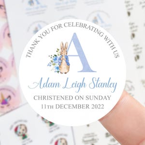 Pode incluir: Adesivo redondo branco com texto "THANK YOU FOR CELEBRATING WITH US" e "Adam Leigh Stanley". Apresenta uma ilustração de Peter Rabbit e uma letra "A" azul. O adesivo é para um batizado em 11 de dezembro de 2022.