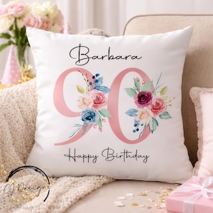 May include: White square cushion with the name "Barbara" and the number "90" in pink, decorated with floral arrangements. The words "Happy Birthday" are written below. The cushion is on a beige sofa.