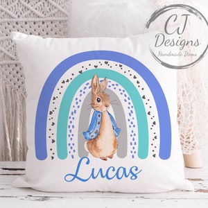 Könnte beinhalten: Ein weißes Kissen mit einem blauen, grünen und grauen Regenbogenmuster. In der Mitte des Regenbogens befindet sich eine Peter Rabbit-Illustration. Der Name "Lucas" ist in Blau unter dem Regenbogen geschrieben.