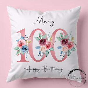 Puede incluir: Cojín blanco con el nombre "Mary" y el número "100" en rosa, adornado con arreglos florales en tonos rosa, azul y burdeos. Las palabras "Happy Birthday" están escritas debajo. Un regalo personalizado.
