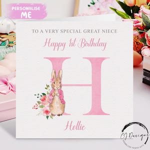 Op de afbeelding: Vierkante verjaardagskaart met de tekst "Happy 1st Birthday" en "To a very special great niece". De roze letter "H" wordt vergezeld door een illustratie van een konijn met een roze jasje. De naam "Hollie" staat eronder.