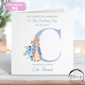 May include: A white greeting card with a watercolor illustration of Peter Rabbit, blue flowers, and a large blue letter "C". Text reads "To a Special Godson, On Your Christening Day, 6th July 2024, Congratulations, Cole Thomas."