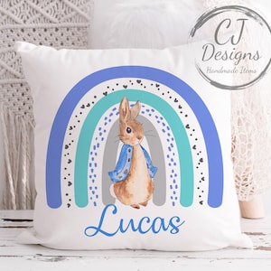 Puede incluir: Cojín cuadrado blanco con una ilustración de acuarela de un conejo con una chaqueta azul frente a un arcoíris azul, turquesa y gris. El nombre "Lucas" está escrito en cursiva azul debajo. El cojín tiene el texto "CJ Designs Handmade Items".