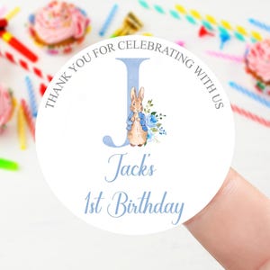 May include: A white circular sticker with the text "Thank you for celebrating with us" and "Jacks 1st Birthday". The letter "J" is in light blue with a Peter Rabbit illustration and floral accents. Cupcakes and party supplies are in the background.