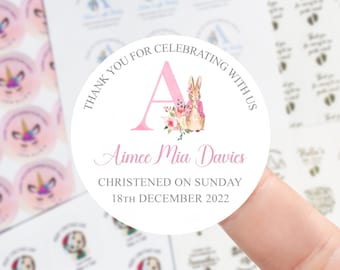 May include: A round white sticker featuring a large pink letter A, a watercolor illustration of a rabbit wearing a pink jacket, and a floral arrangement. The text reads: THANK YOU FOR CELEBRATING WITH US, Aimee Mia Davies, CHRISTENED ON SUNDAY 18TH DECEMBER 2022.