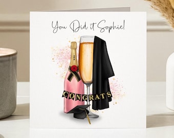 May include: A graduation greeting card with a pink champagne bottle, a filled champagne glass, and a black graduation gown. The card reads "You Did it Sophie!" and "Congrats" in gold letters. A small graduation cap is also present.