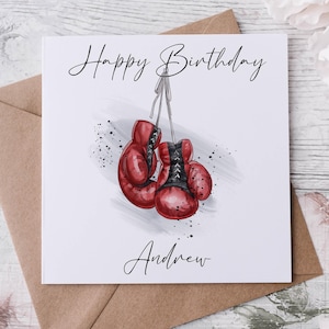 May include: A birthday card with the words "Happy Birthday Andrew" in script. It features a watercolour illustration of red boxing gloves with black detailing, suspended by a string, and a splatter effect.