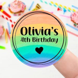 May include: A circular sticker with a rainbow gradient, displaying the text "Olivia's 4th Birthday" in black, and a black heart. Cupcakes, candles, and party decorations are visible in the background.