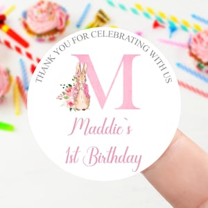 Peut inclure: Un autocollant rond blanc avec une grande lettre M rose, une illustration aquarelle d'un lapin avec des fleurs roses, et le texte THANK YOU FOR CELEBRATING WITH US, Maddie's 1st Birthday en lettres roses cursives et capitales.