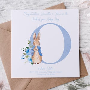 Könnte beinhalten: Babykarten-Design mit Peter Rabbit in blauem Jackett neben einem blauen "O". Auf der Karte steht "Herzlichen Glückwunsch Samantha & James zur Geburt Ihres Baby Boy". Sie enthält auch den Namen, das Geburtsdatum, die Uhrzeit und das Gewicht des Babys.