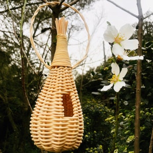Peut inclure: Un nichoir en osier tressé avec une ouverture ronde, suspendu à une branche avec des fleurs blanches.
