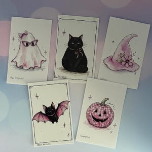 May include: Five rectangular art prints featuring illustrations in pink and black ink. The images depict a ghost with glasses, a black cat, a pink witch hat with flowers, a bat with pink wings, and a pink pumpkin, each labeled with handwritten text.