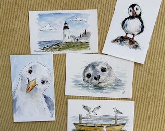 May include: Five rectangular watercolor art prints featuring coastal themes: a lighthouse labeled Pemaquid Point Lighthouse, a puffin on a rock, a close-up of a seagull, a seal head emerging from water, and a yellow boat with seagulls labeled Lunch with a View.