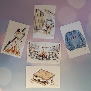 May include: Five rectangular art prints featuring watercolor illustrations: a toasted marshmallow on a stick, a wooden chair with a mug, a blue sweatshirt with small icons, a campfire between two wooden chairs with string lights, and a s'more.