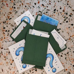 May include: A collection of dark green paper envelopes, some containing white cards with blue watercolor question mark illustrations and decorative accents. The envelopes are arranged in a scattered, overlapping pattern, showcasing various hand-drawn designs on the visible card edges.